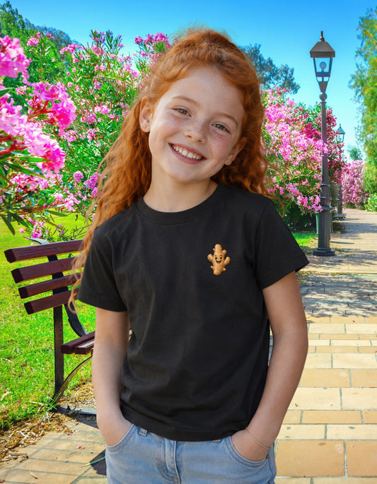 A smiling young girl with red hair stands outdoors in a park-like setting with pink flowers, a bench, and a lamp post, wearing a black short-sleeve T-shirt with a small tan bear graphic on the left chest and light blue jeans.