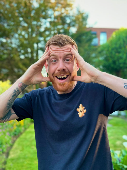 Man with tattoos on arms wearing a blue t-shirt with a logo, outdoors with greenery.