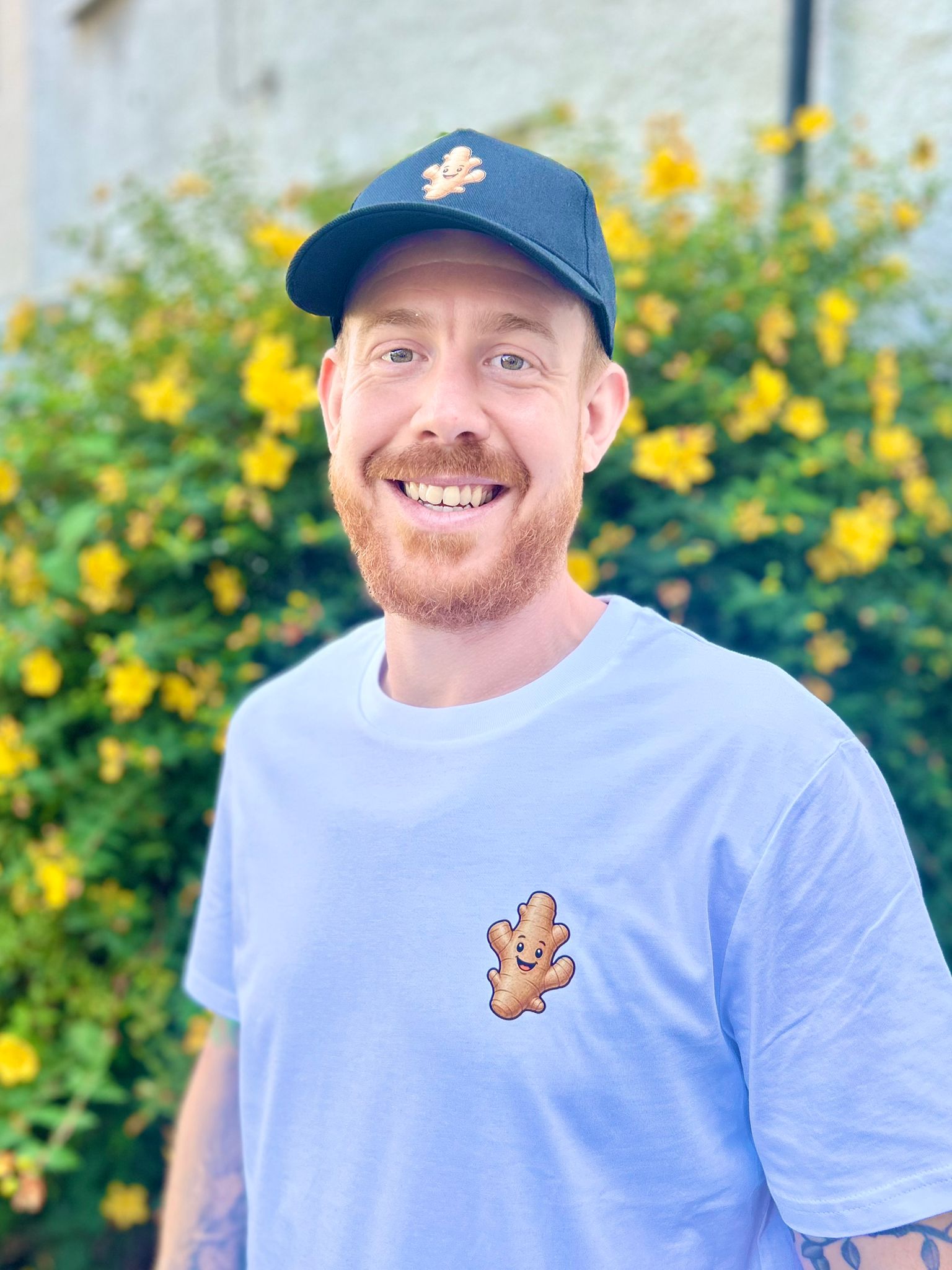 Man wearing a blue cap and light blue t-shirt with a cartoon character, standing in front of yellow flowers.