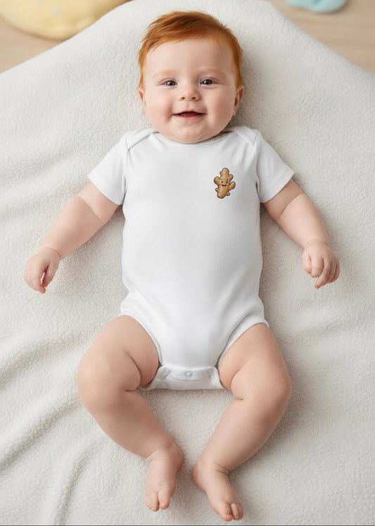 A smiling baby with red hair lies on a white blanket, wearing a white short-sleeve bodysuit with a small brown teddy bear graphic on the left chest.